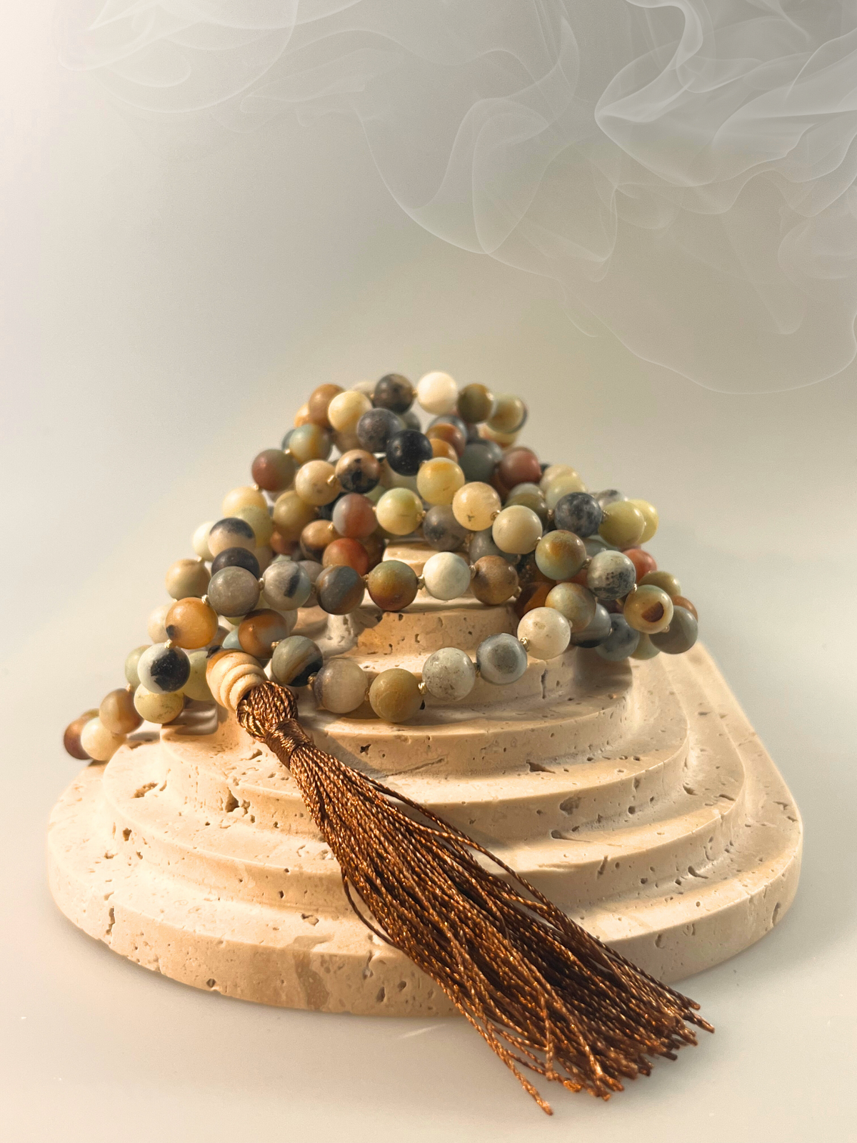 Eine handgeknüpfte Mala-Kette aus matten Amazonit-Perlen in sanften Naturtönen von Grün, Grau, Beige und Rotbraun. Die Kette ist spiralförmig gelegt und mit einer dunkel schimmernden Quaste sowie einer hellen Holz-Guru-Perle versehen. Das Schmuckstück ist freigestellt vor neutralem Hintergrund dargestellt und eignet sich ideal für Meditation, Erdung und Momente der inneren Balance.