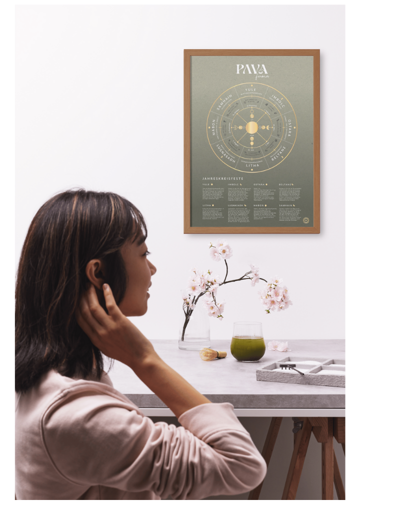 Frau sitzt entspannt mit einer Schale Matcha inmitten von Blumen, während an der Wand das stilvolle PAVA Jahreskreis-Poster mit goldener Schrift und natürlichen Farben hängt. Eine harmonische Szene voller Ruhe und spiritueller Inspiration.