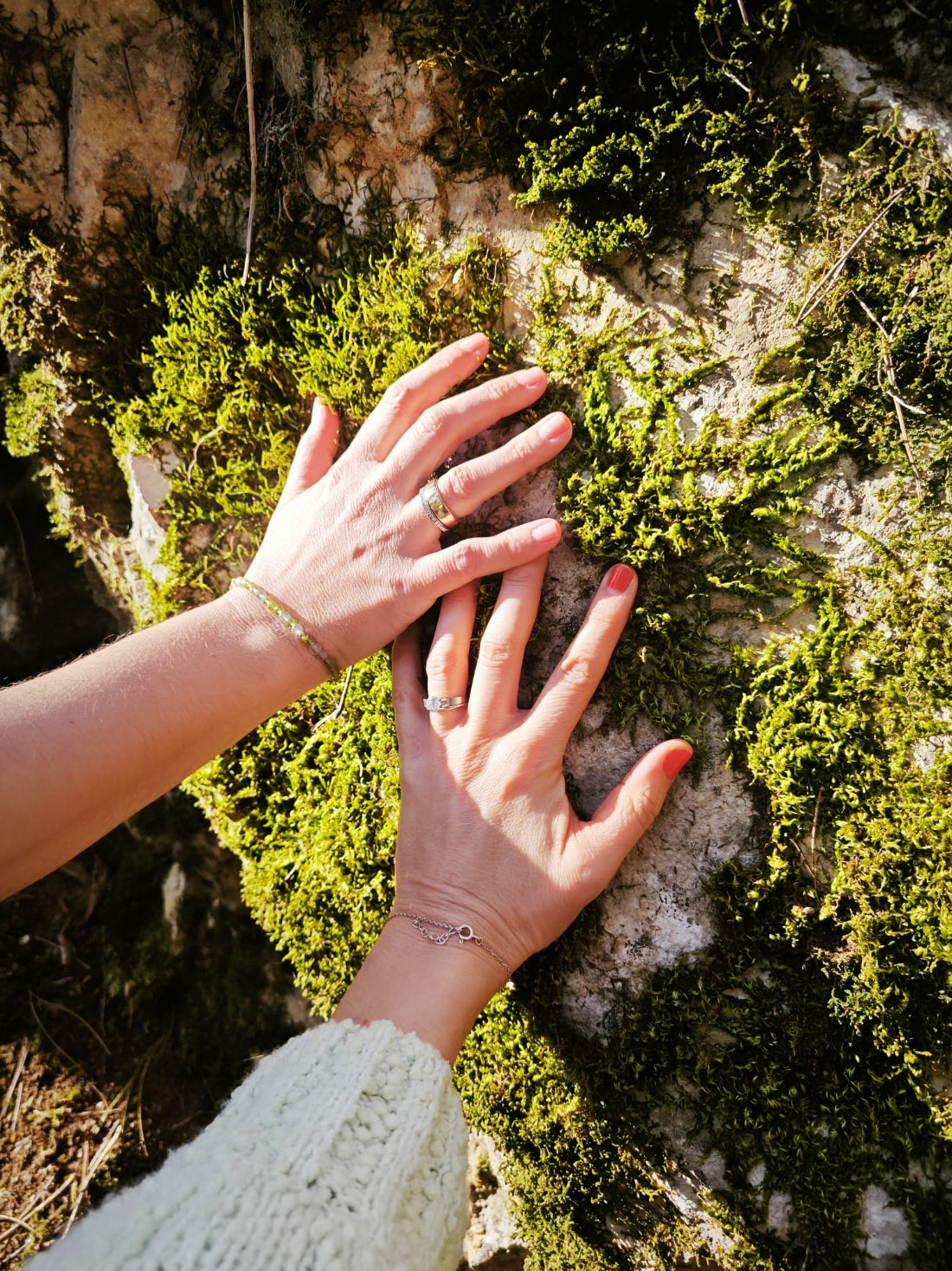 Zwei Hände berühren sanft einen moosbewachsenen Felsen in der Sonne – ein Moment tiefer Naturverbindung. Zarte Armbänder und Ringe unterstreichen die feminine Energie. Ein Bild für Erdung, Achtsamkeit und das bewusste Spüren der Elemente.