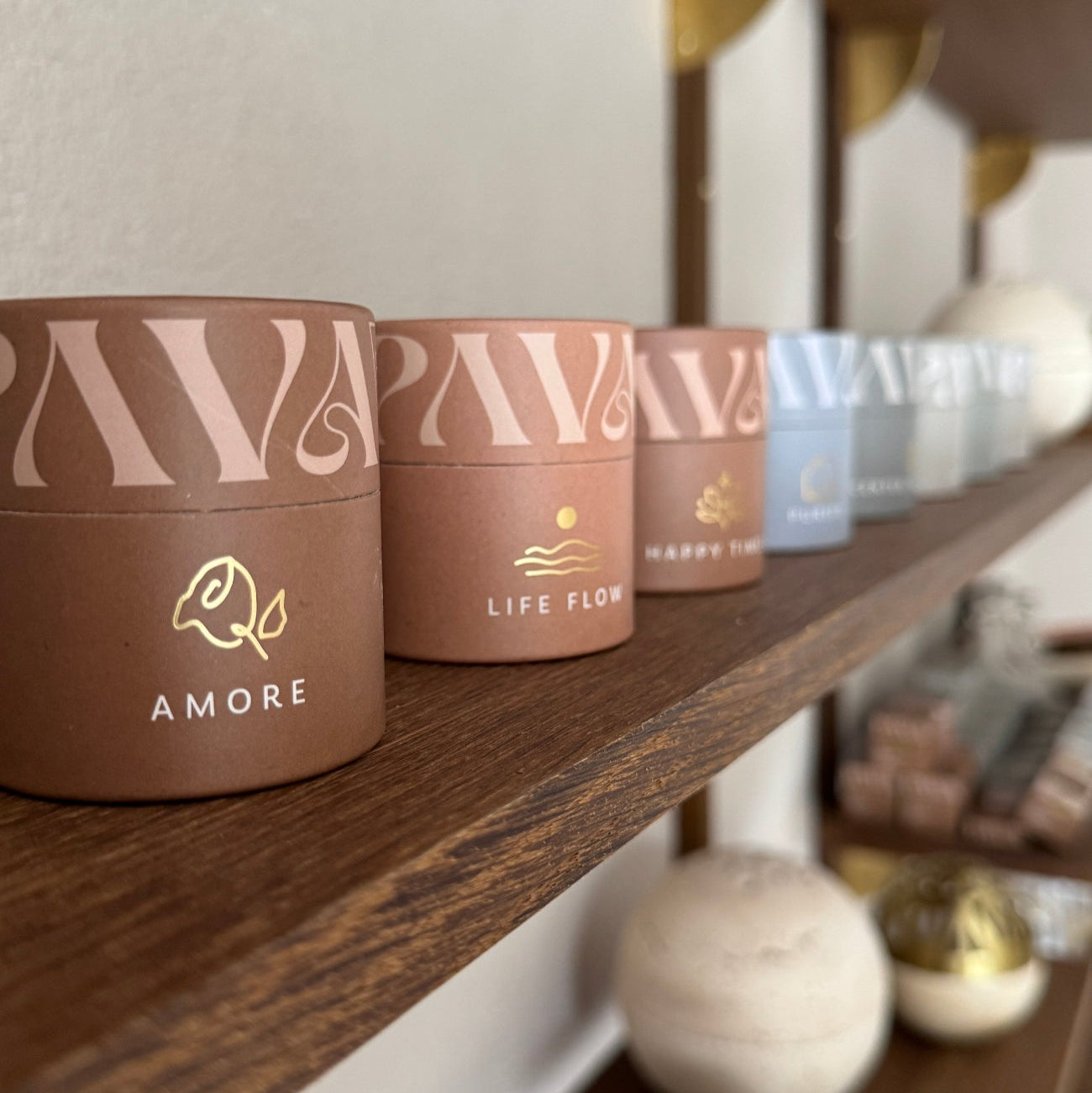 Nahaufnahme eines Holzregals mit den Duftmischungen von PAVA FUMA in farblich abgestimmten Design-Tuben. Im Fokus stehen die Sorten „Amore“, „Life Flow“ und „Happy Times“ in warmen Naturtönen mit goldgeprägtem Logo. Die Produkte sind sorgfältig arrangiert, im Hintergrund folgen weitere Sorten in zarten Blau- und Cremetönen. Das Bild vermittelt Ästhetik, Duftvielfalt und die hochwertige Markenwelt von PAVA FUMA – inspiriert von Emotion, Natur und Ritual.