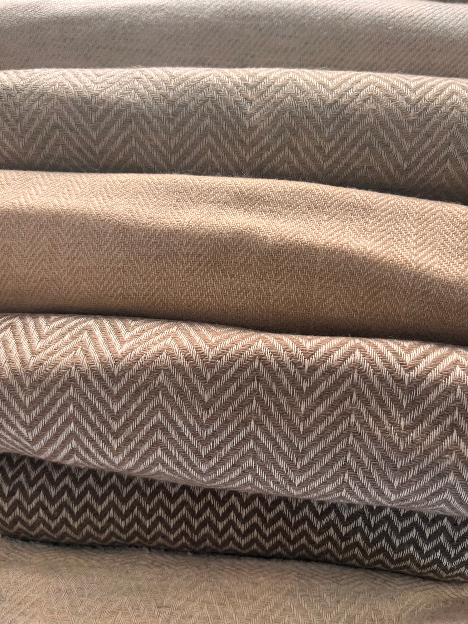 Detailaufnahme von mehreren handgewebten Kamelhaarschals in Naturtönen – elegant gestapelt und mit feinem Fischgrätmuster in variierenden Braunnuancen. Die natürliche Struktur und das edle Finish unterstreichen die hochwertige Qualität der fair produzierten, nachhaltigen Accessoires. Ideal für Interior-Liebhaber*innen mit Sinn für Ästhetik und Wärme.