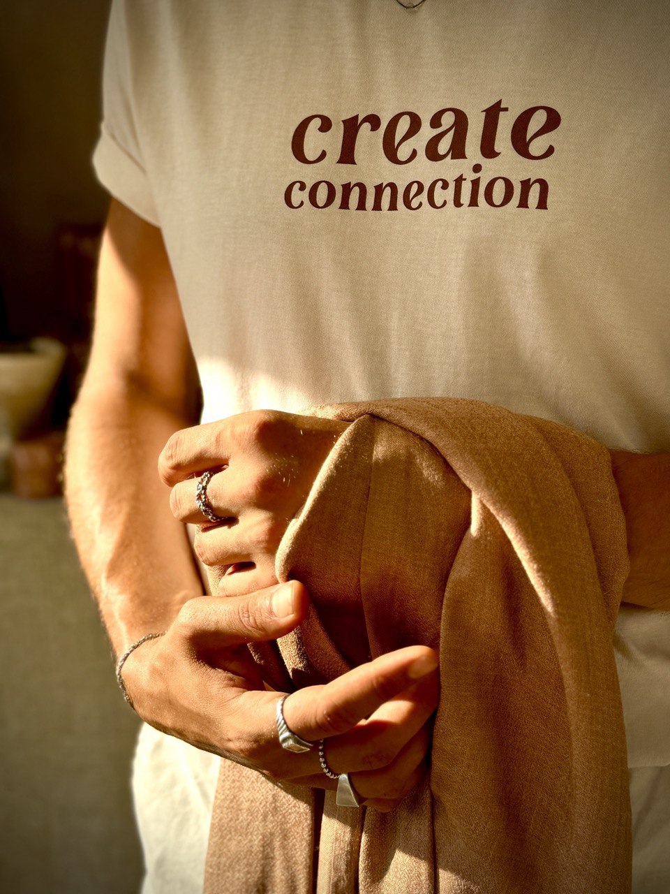 Sonnenlicht fällt auf ein cremefarbenes T-Shirt mit der Aufschrift „create connection“. Eine Hand hält einen weichen, naturfarbenen Schal – ein Moment der Achtsamkeit, Stil und Verbindung. Schmuckdetails unterstreichen die individuelle Note.