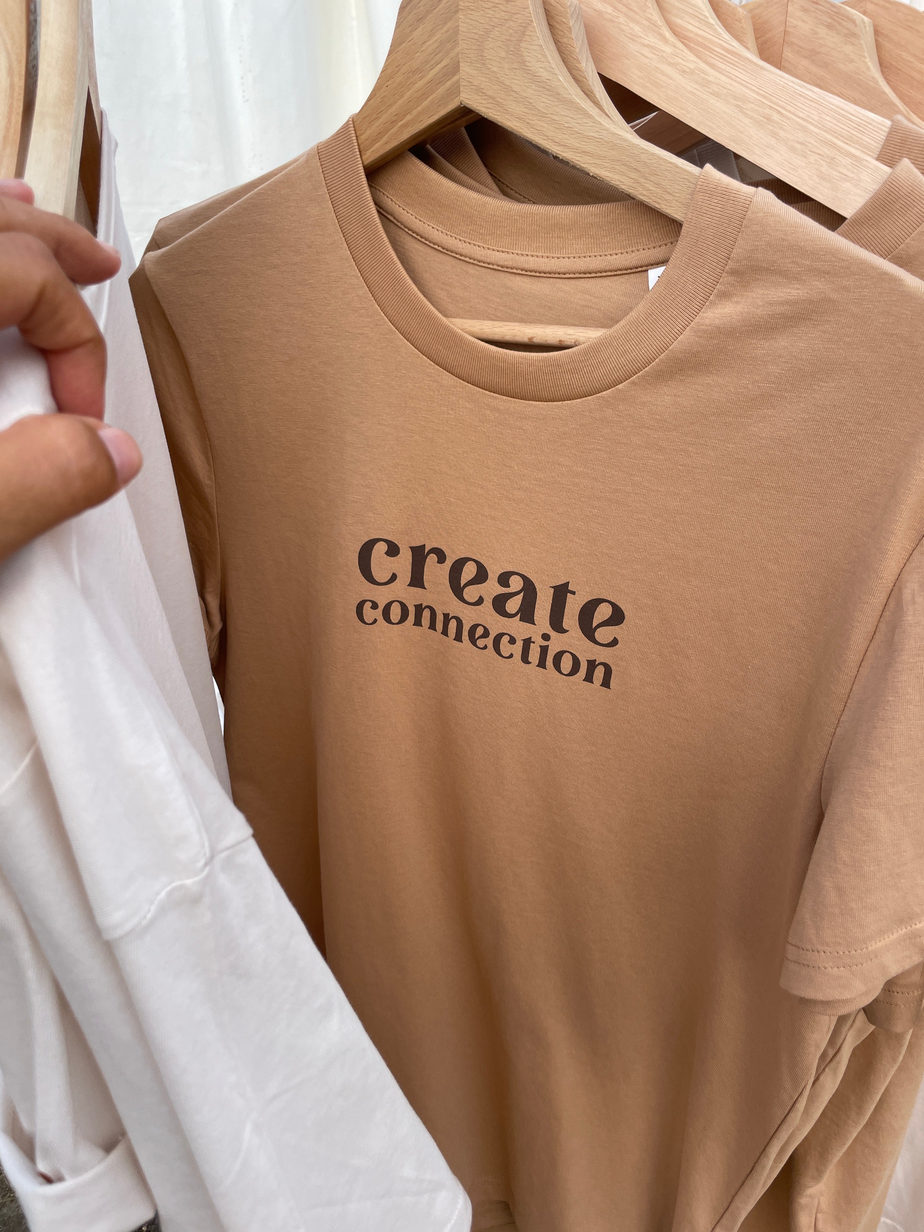 Beigefarbenes T-Shirt mit dem Statement „create connection“ in stilvoller Typografie – auf einem Holzkleiderbügel zwischen weiteren natürlichen Textilien. Reduziert, bewusst und modern – ein Ausdruck von Verbundenheit und Stil.