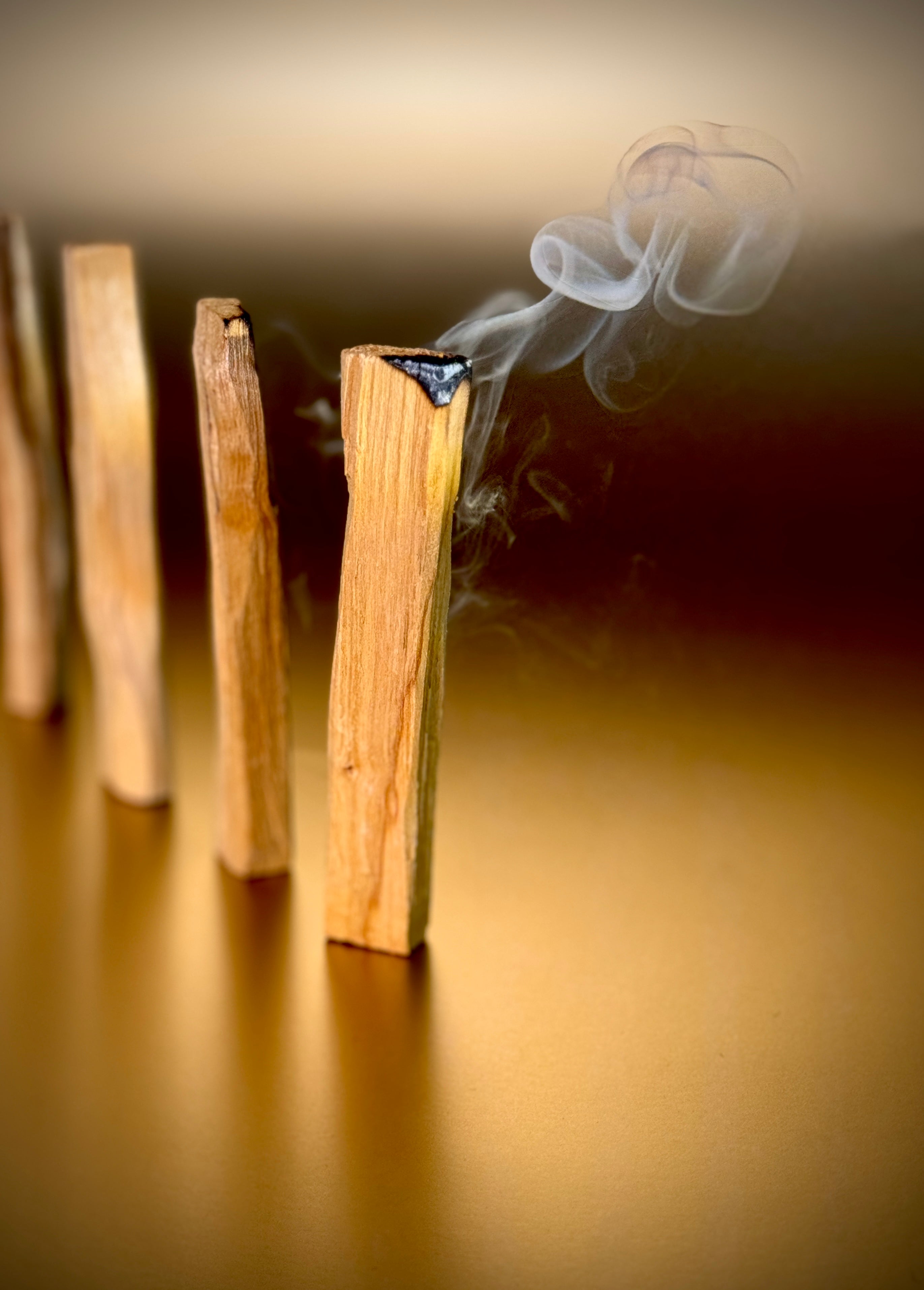 Auf diesem Bild siehst du mehrere Palo Santo Hölzer aufgereiht auf einem goldenen Hintergrund. Das erste Palo Santo Holz glimmt und räuchert. Die Farben sind warm und harmonisch