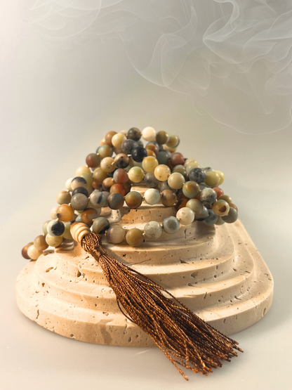 Eine handgeknüpfte Mala-Kette aus matten Amazonit-Perlen in sanften Naturtönen von Grün, Grau, Beige und Rotbraun. Die Kette ist spiralförmig gelegt und mit einer dunkel schimmernden Quaste sowie einer hellen Holz-Guru-Perle versehen. Das Schmuckstück ist freigestellt vor neutralem Hintergrund dargestellt und eignet sich ideal für Meditation, Erdung und Momente der inneren Balance.