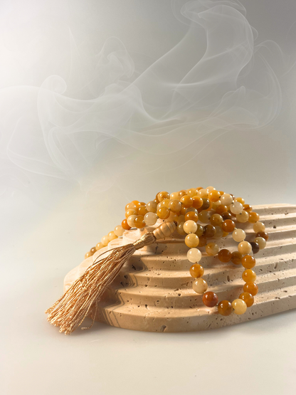 Eine handgeknüpfte Mala-Kette aus polierten Honigcalcit-Perlen in warmen Gelb- und Goldtönen, spiralförmig gelegt und mit einer goldschimmernden Quaste sowie einer glatten Guru-Perle aus hellem Holz veredelt. Das Schmuckstück ist freigestellt auf hellem Hintergrund dargestellt und eignet sich ideal für Meditation, spirituelle Rituale oder als energetisches Accessoire mit sonniger Ausstrahlung.