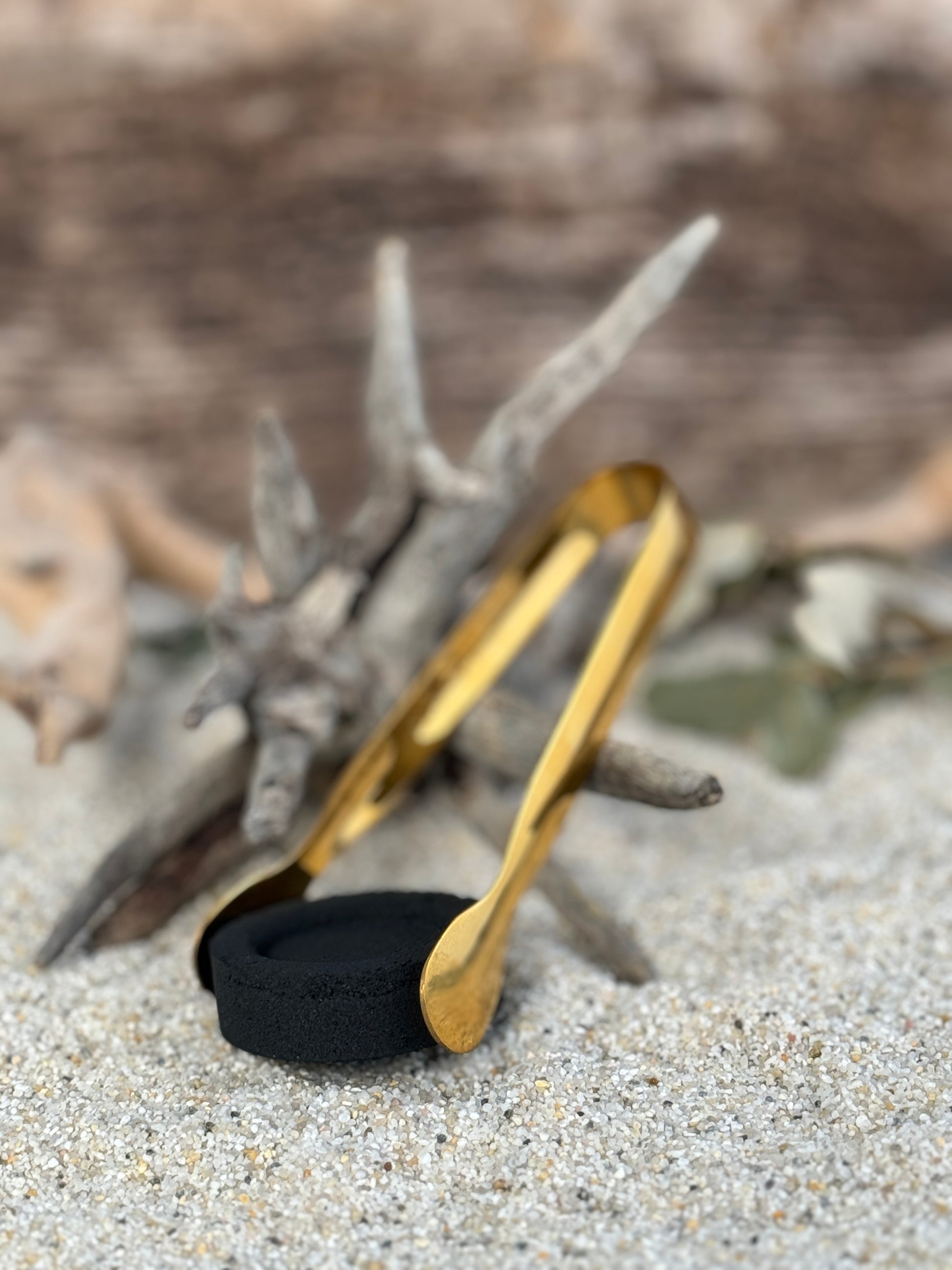 Räucherzange in gold mit einer Räucherkohle auf einem Sandbett an ein Treibholz angelehnt