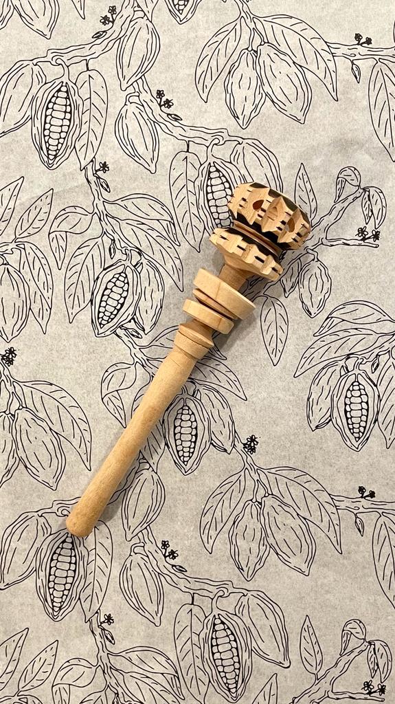 Handgedrehter Molinillo aus Holz – traditioneller Rührstab zur Zubereitung von zeremoniellem Kakao – liegt auf einem gezeichneten Hintergrund mit Kakaopflanzen-Motiv. Symbol für Genuss, Achtsamkeit und kulturelle Verbindung.