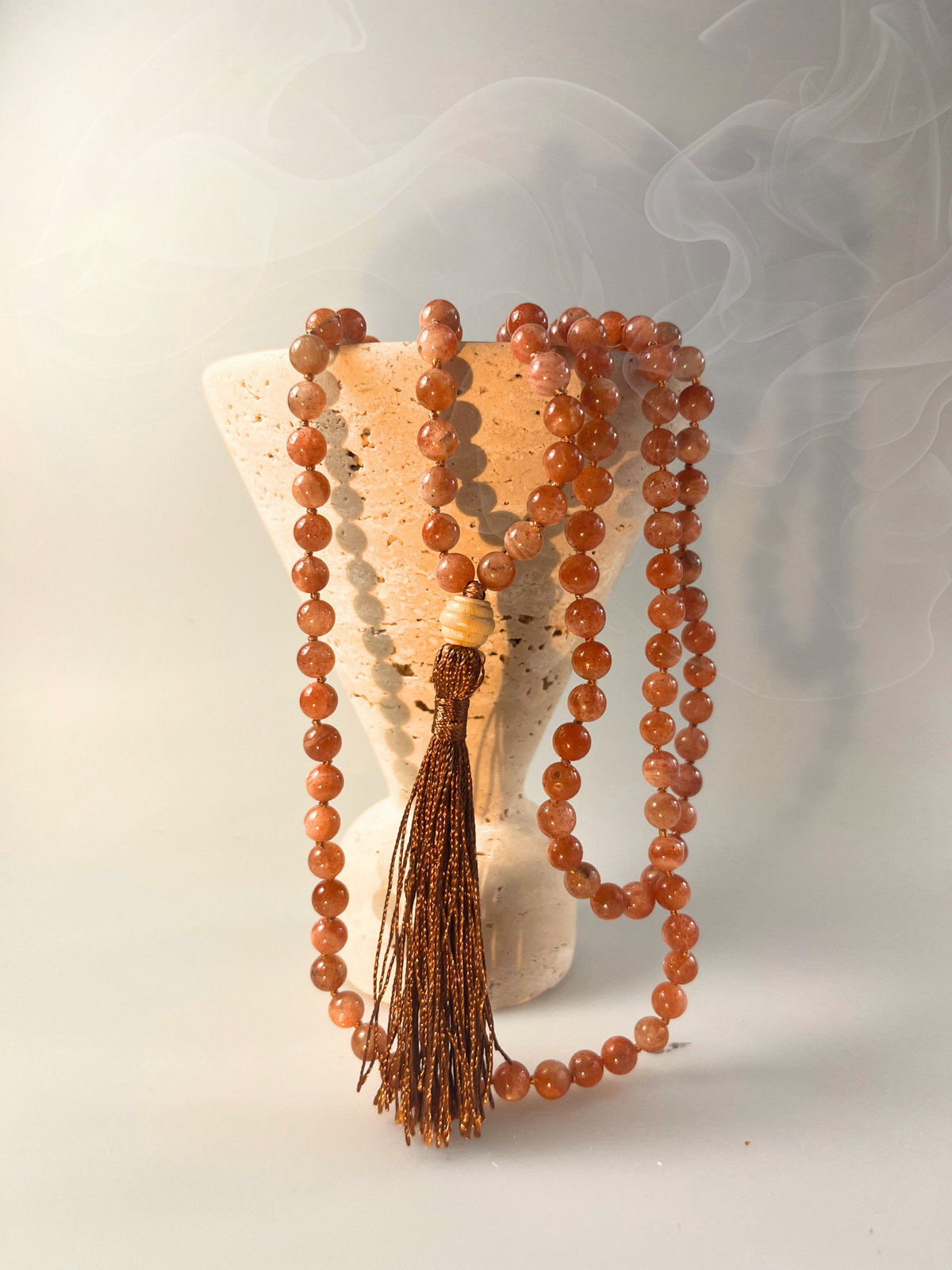 Handgeknüpfte Mala-Kette aus Sonnenstein-Perlen in warmen Kupfer- und Rosétönen, arrangiert in weicher Schlaufenform. Die Kette ist mit einer schimmernden dunkelgoldenen Quaste und einer hellen Holz-Guru-Perle veredelt. Das Schmuckstück ist freigestellt vor transparentem Hintergrund dargestellt und eignet sich ideal für energetisierende Rituale, Meditation und als stilvolles Accessoire.